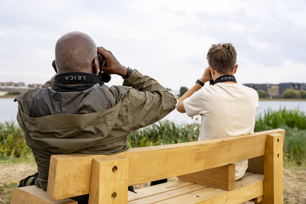 Two London Wildlife Trust staff members sitting on a bench looking out over Walthamstow Wetlands using Leica binoculars