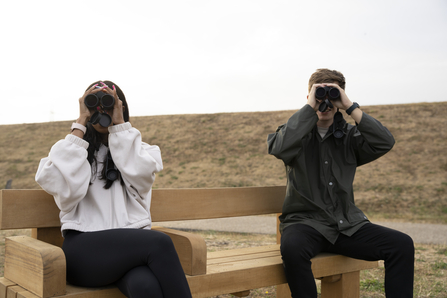 Two people sit on a bench using binoculars to birdwatch