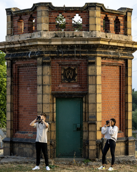 Two people stand in front of a ornate brick building with a green door, looking through binoculars