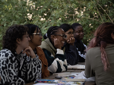 A group of young adults at an outdoor table surrounded by trees, with paper and coloured pens on the table. Smiling during a creative writing workshop.