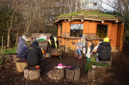 A group of people sat on short log stumps in front of a round orange house with a grassy roof