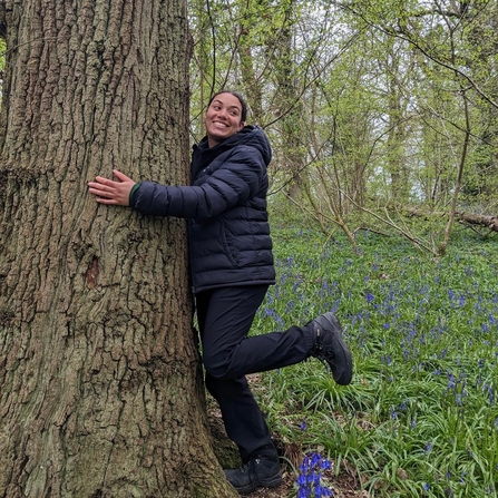 Keeping it Wild trainee Yasmin hugging a tree whilst smiling in a woodland with blue bells on the ground.