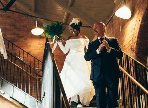 Two people at a wedding walking down the stairs, one wears a wedding dress holding a bouquet and the other wears a suit