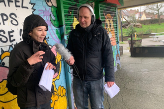 A man holding a microphone interviewing a woman outside in front of a colourful, graffitied wall