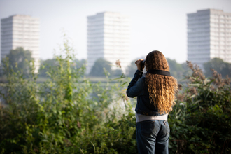 Young woman with binoculars looking across the water, tower blocks in background. She has her back to the camera