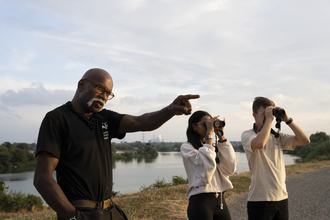 Two people looking through binoculars while a third person points, behind them is a reservoir at Walthamstow Wetlands