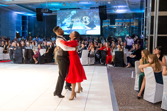 Two people dance in front of an audience of people, one wears a red dress the other a suit