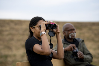 Two people sit on a bench, one uses binoculars to birdwatch the other sits and smiles