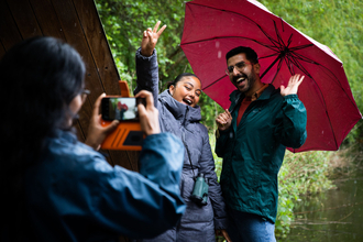 Two people pose in front of a camera one holds an umbrella and the other poses with their hands in the air another person stands in front of the camera taking a picture