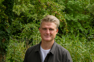 A man standing in front of a green backdrop full of vegetation. The person pictured is David Mooney