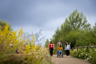 Four people making their way down a path surrounded by vegetation, the person in the middle pushes someone in a wheelchair and the two others walk either side