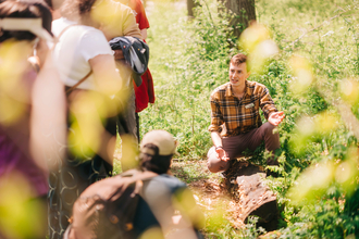 Young man delivering a walking tour crouched down pointing at a log for a crowd to observe