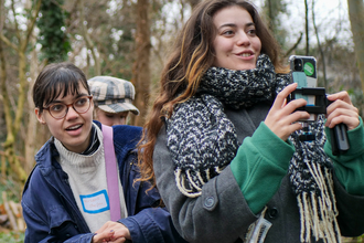 Two young people outdoors in a wooded area, one holding a phone, both dressed warmly in coats and scarves