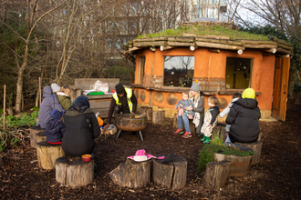 A group of adults and infants sit outdoors in a log circle around a firepit