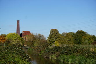 A tall brown chimney and red tiled triangular roof are visible above a line of trees in varying shades of green. A river runs through the image from the left, and blue sky fills the background.