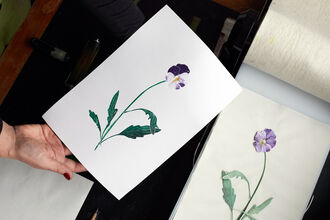 A print of a lilac & purple flower with yellow stamen and green leafy stem on a white sheet of paper. The etched version of this flower made from Tetrapak, that was used to make the print, sits to the right. Its colours are the same but more muted. 