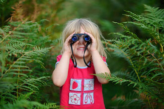 A child looks through binoculars towards the camera, surrounded by ferns and greenery