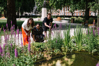 Three people smiling and looking at a small pond surrounded by purple flowers in a sunny park.