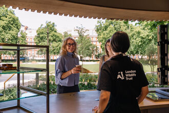 Two women standing at a kiosk with coffee, green garden in the background