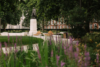 A large statue of U.S. President Franklin Roosevelt stands on a plinth in the background. A white footpath curves past it from the foreground, with green and purple plants out of focus at the front of the frame.