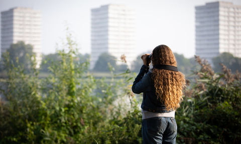 Young woman with binoculars looking across the water, tower blocks in background. She has her back to the camera