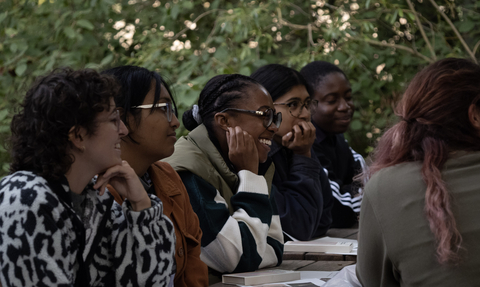 A group of young adults at an outdoor table surrounded by trees, with paper and coloured pens on the table. Smiling during a creative writing workshop.