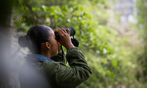 A young woman in a green jacket looking through binoculars into the trees