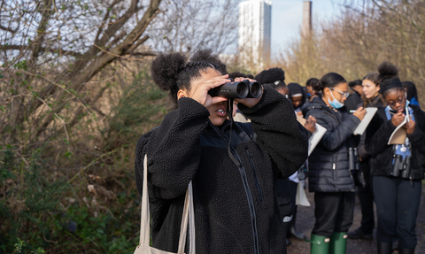 Group of people walking through Walthamstow Wetlands, person at front holding binoculars up to their eyes