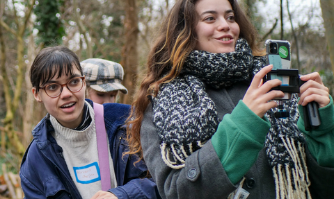 Two young people outdoors in a wooded area, one holding a phone, both dressed warmly in coats and scarves
