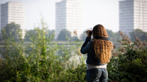 Young woman with binoculars looking across the water, tower blocks in background. She has her back to the camera