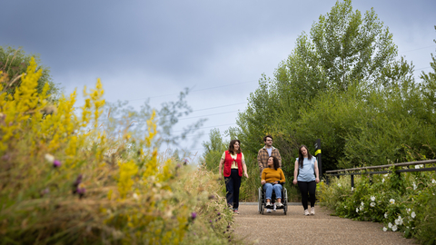 Four people making their way down a path surrounded by vegetation, the person in the middle pushes someone in a wheelchair and the two others walk either side