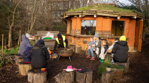A group of adults and infants sit outdoors in a log circle around a firepit