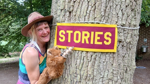 Woman stood next to a tree holding a fox puppet pointing at a stories sign