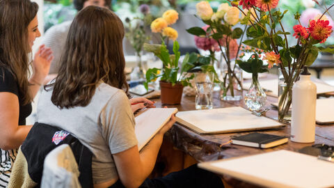 Two people with drawing boards sit in front of vases of pink, yellow & white flowers