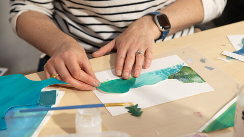 A person's hands are shown placing various shapes in different shades of green to create a collage of a landscape.