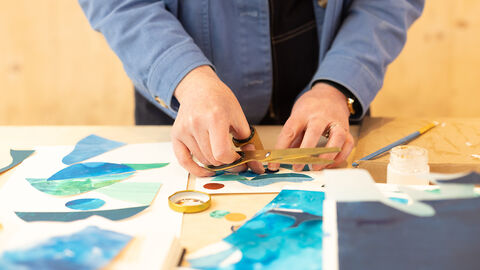A person's hands are shown holding golden scissors whilst placing paper of varying colours onto a sheet to make a collage.