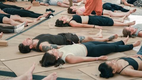 A group of people in exercise clothing lie flat on yoga mats whilst a teacher in baggy orange trousers and black vest walks between them.