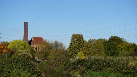 A tall brown chimney and red tiled triangular roof are visible above a line of trees in varying shades of green. A river runs through the image from the left, and blue sky fills the background.