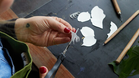 An over-the-shoulder view of a person using scissors to cut the shape of some petals out of a piece of white Tetrapak. Five leaf-shaped cutouts sit on the table below. 