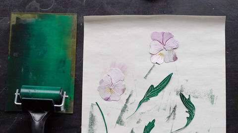 Individual cutouts of lilac petals and green leaves are arranged on a white piece of paper. There is a green paint roller to their left, and green smudges on the paper where the roller has been used.