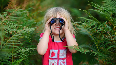A child looks through binoculars towards the camera, surrounded by ferns and greenery
