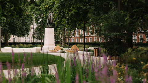 A large statue of U.S. President Franklin Roosevelt stands on a plinth in the background. A white footpath curves past it from the foreground, with green and purple plants out of focus at the front of the frame.