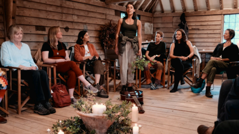 A person stands in the middle of a group of people, inside a wood-panelled room. They are performing a poem. In the centre of the room is a selection of branches and candles. 