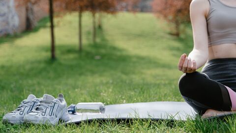 A woman in sportswear meditating on a yoga mat outdoors with shoes and water bottle nearby.