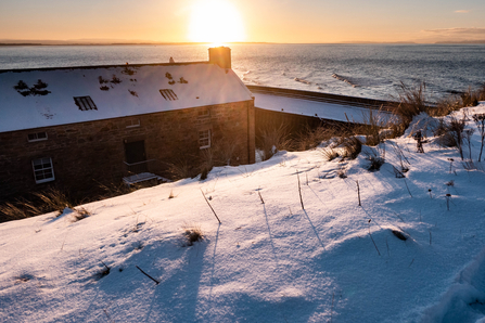 A landscape filled with snow, a bank of snow in the foreground, two buildings and the horizon and low sun behind. 