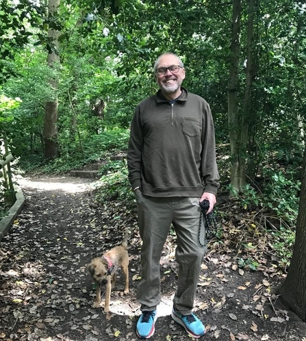 Image of a man stood in a woodland on a mud path with his small dog next to him, trees surrounding them