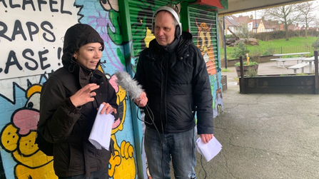 A man holding a microphone interviewing a woman outside in front of a colourful, graffitied wall