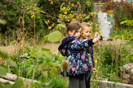 Two children stand pointing at something off camera, surrounded by vegetation.