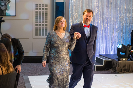 Two people walk holding hands wearing a sparkly silver dress and a navy blue suit with a red bow tie