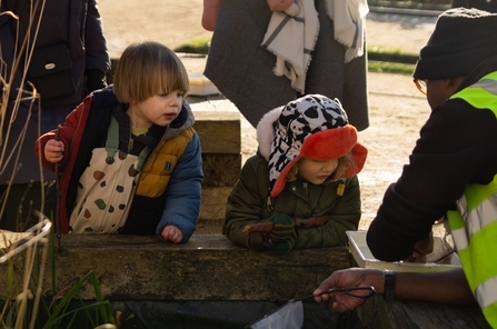 Two children lean on a wooden ledge of a pond looking in a white tray and person is pointing at 